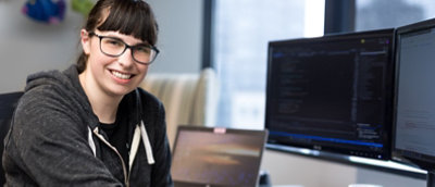 A smiling young woman with glasses sits at a desk with multiple computer screens displaying code.