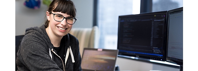 A smiling young woman with glasses sits at a desk with multiple computer screens displaying code.