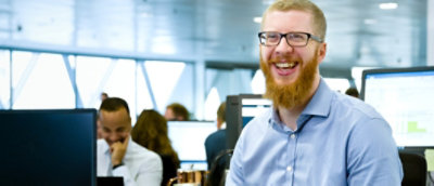 Smiling man with beard in office environment, sitting in front of a computer screen, other workers in background.