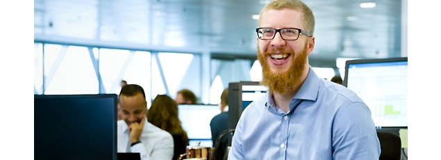 Smiling man with beard in office environment, sitting in front of a computer screen, other workers in background.