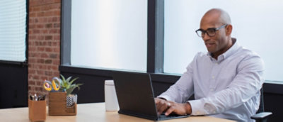 A bald man wearing glasses and a striped shirt works on a laptop in a modern office with brick walls and a window.