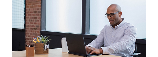 A bald man wearing glasses and a striped shirt works on a laptop in a modern office with brick walls and a window.