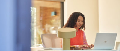 A woman working on a laptop at a home office desk, bathed in natural light from a nearby window.