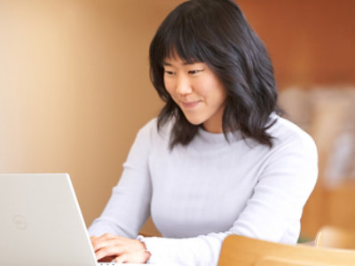 A young woman with black hair, wearing a light turtleneck sweater, is focused on working on her laptop