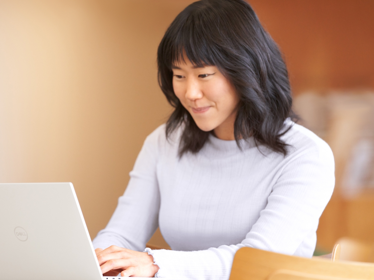 A young woman with black hair, wearing a light turtleneck sweater, is focused on working on her laptop