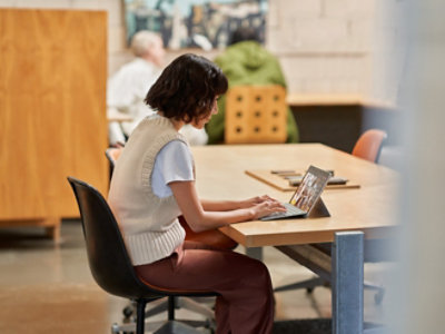Woman using a laptop at a library table, viewed from the side, with other people in the background.