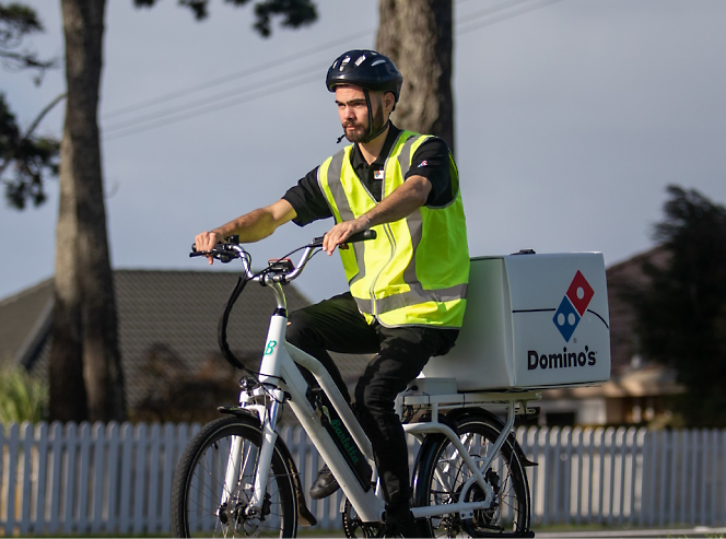 A domino's delivery person wearing a helmet and reflective vest rides an electric bike 
