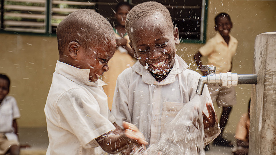 Two children joyfully splash water from an outdoor faucet.
