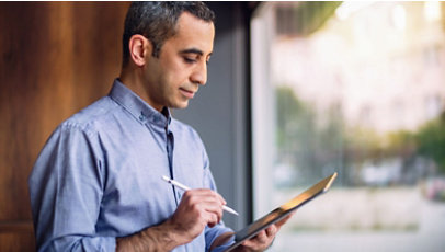 A person in a blue shirt is standing indoors, holding a pen and a tablet.