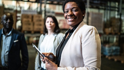 Three people in a warehouse, one holding a tablet near shelves of boxes.