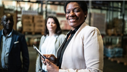 Three people in a warehouse, one holding a tablet near shelves of boxes.