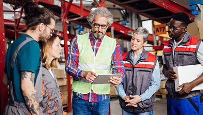 Four warehouse workers, one with a tablet and another with a clipboard, in an aisle with shelves and boxes.