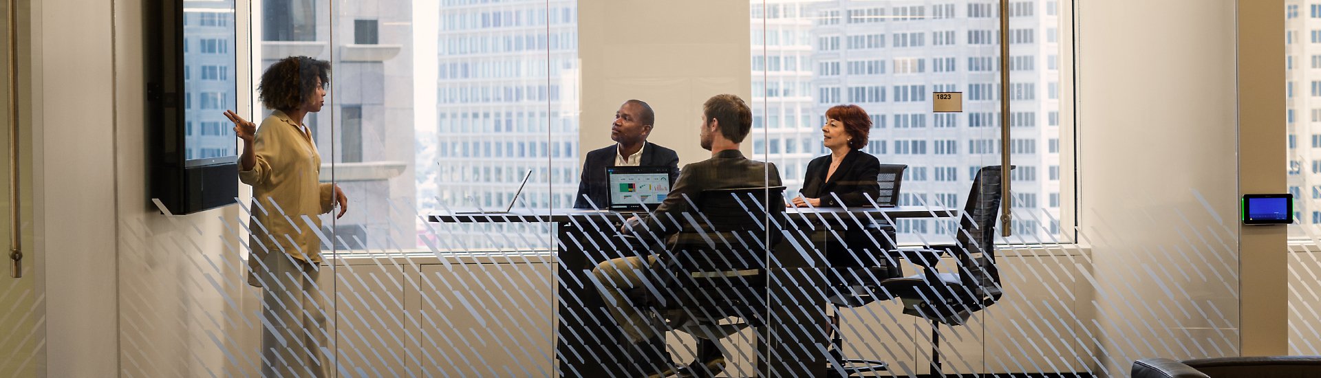 Three people seated at a table in a modern conference room watching a presentation being made by a fourth person who is standing at a large, wall-mounted display.