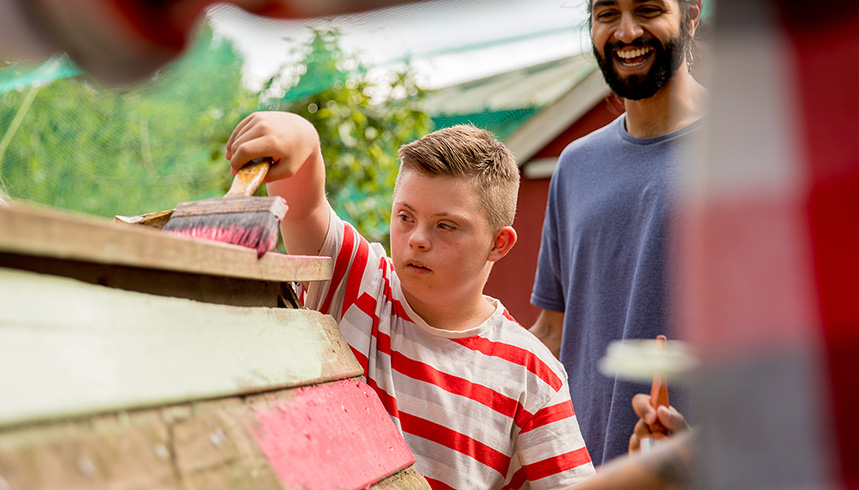 A young boy painting a wooden structure outdoors with an adult nearby.
