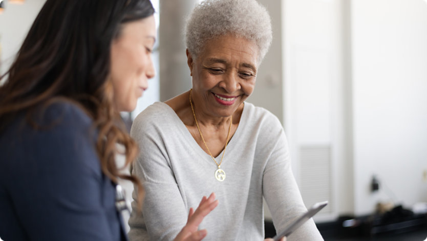 An older woman smiles while looking at a tablet with another woman indoors.