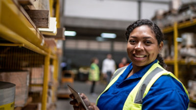 A smiling woman in a warehouse holding a tablet.