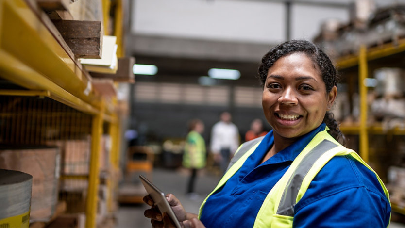 A smiling woman in a warehouse holding a tablet.
