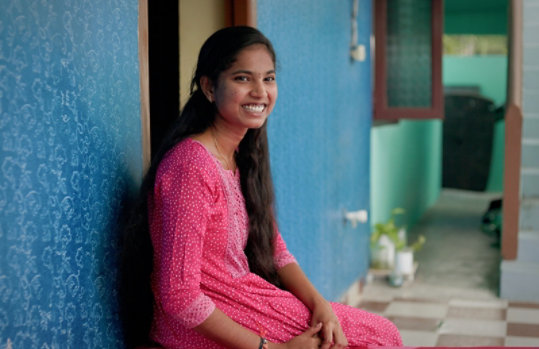 A young woman smiling while sitting outside a door.