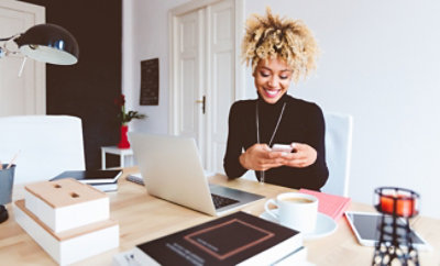 Osoba kovrdžave kose sedi za stolom na kojem se nalaze laptop, telefon, beležnice i šolja kafe,