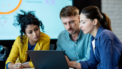 Three colleagues collaborate on a laptop in an office setting.