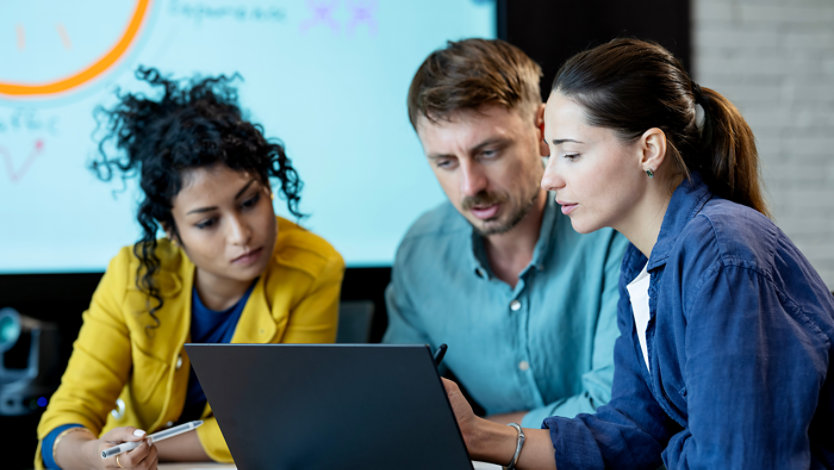 Three colleagues collaborate on a laptop in an office setting.