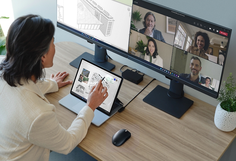 Persona che usa un tablet per presentare progetti relativi all'architettura durante una riunione di Teams visualizzata su due monitor desktop.