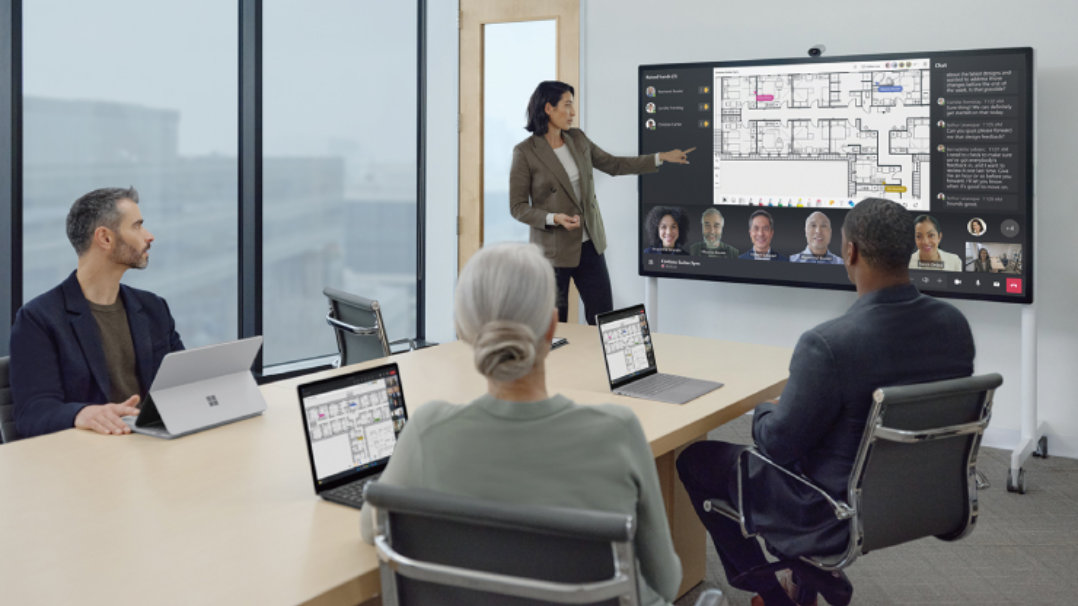 Surface Hub 3でTeamsの会議に参加し、女性が立って説明し2人の男性と1人の女性が聞いている