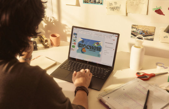 Eine Person arbeitet mit einem Surface Laptop an einem Schreibtisch, der mit Notizbüchern, handschriftlichen Notizen und Schreibtischutensilien übersät ist.