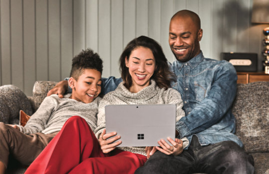 A family, sitting on a couch, using a Surface device together.