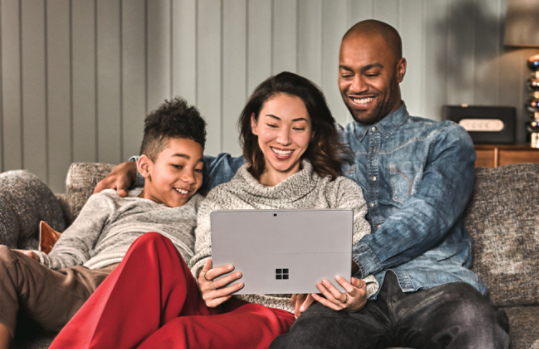 A family, sitting on a couch, using a Surface laptop together. 