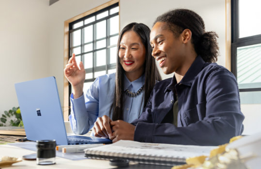 Two people sit together at a table using a Sapphire Surface Laptop in a bright room.