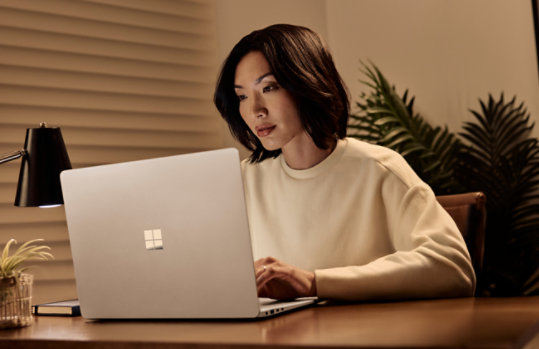 A woman works on a silver Surface laptop at a desk in a warmly lit room.