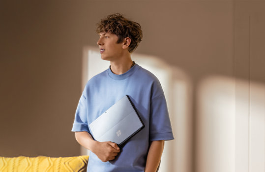 Person standing in a room beside a sofa, holding a Surface Pro in their arm.