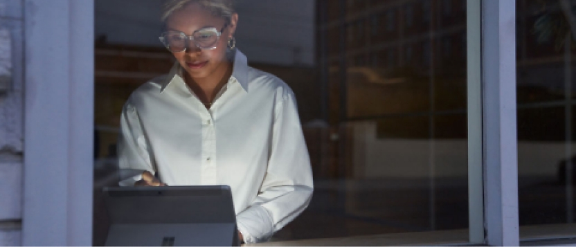 Uma mulher de óculos está a utilizar um computador tablet em frente a uma janela.