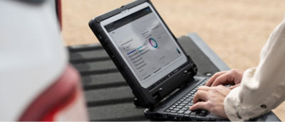 Person typing on a rugged laptop placed on the back of a vehicle, with a screen displaying a dashboard featuring charts and data.