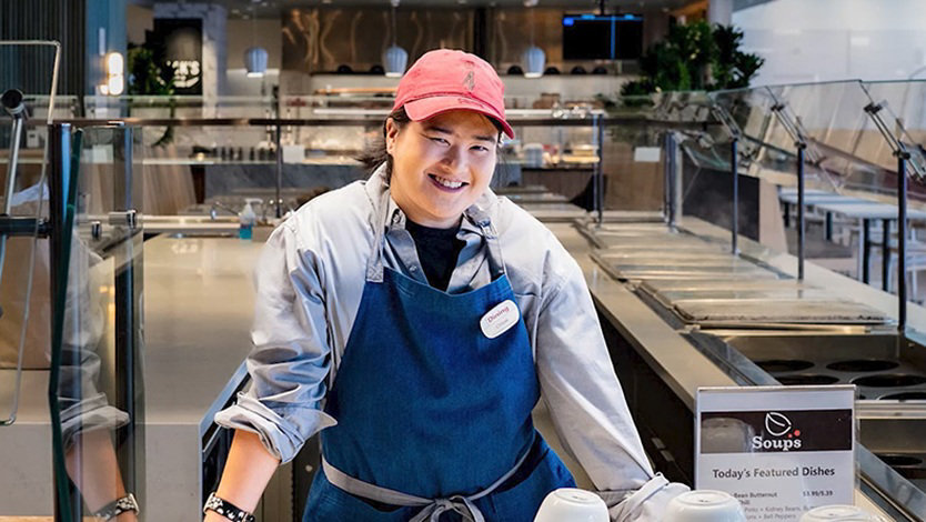 Chloe is smiling standing behind the counter of a cafe at Microsoft. She is wearing a red ball cap and a blue shirt and blue apron.