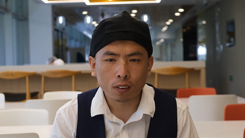 Hongqiang is seated wearing a white shirt and black waistcoat. He is in a cafe in a Microsoft building, with white and oranges chairs in the background.