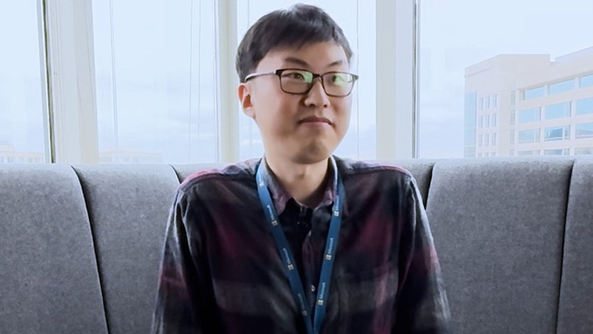 Minkee is seated wearing a plaid flannel shirt. He is smiling, wearing glasses. He is seated on a high-backed couch in an office setting with large windows.