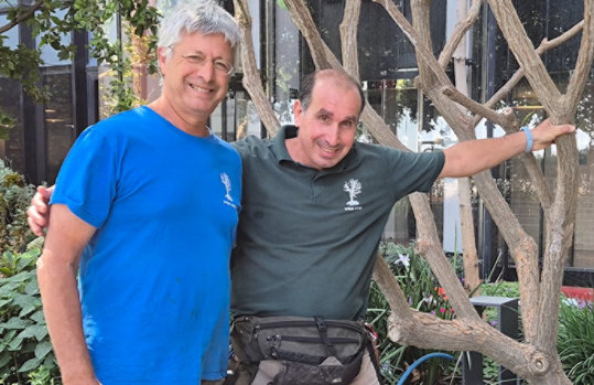 Two men in t-shirts stand together shoulder to shoulder, smiling. They stand outdoors with a tree and other landscaping behind.