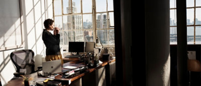 אדם עומד ליד חלונות משרד, מביט החוצה ולוגם מספל. המשרד מלא בשולחנות, מחשבים