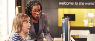 Ženska z dolgimi lasmi, ki nosi očala, stoji v sobi, kjer je vidno besedilo »dobrodošli v svet«.