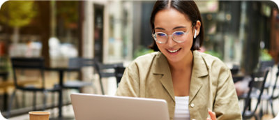 A person with glasses and earphones sits at an outdoor cafe, smiling at a laptop screen.