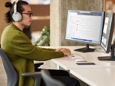 Personne portant des lunettes et des écouteurs, travaillant sur un ordinateur de bureau avec plusieurs écrans
