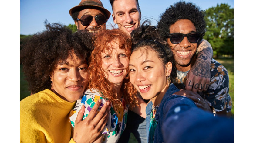 Six diverse young friends smiling closely together for a selfie in a sunlit park, radiating warmth and belonging.