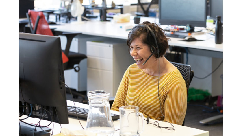 A person in a yellow sweater wearing a phone headset laughs while working at her computer in a bright, open-plan office.