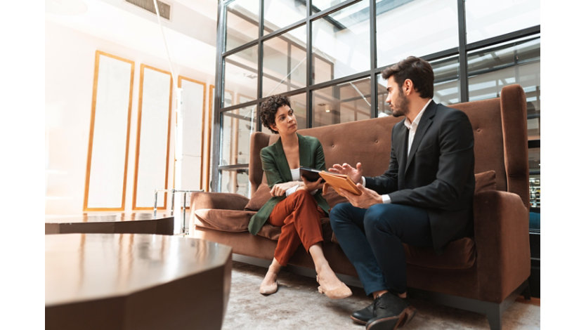 Two colleagues seated on a sofa in a modern office discuss work while reviewing information on tablet devices. One person gestures as they speak, while the other listens attentively.
