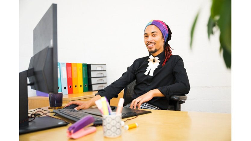 Person seated in an office setting, smiling toward the camera. They are wearing a black top, a colorful head wrap, and a decorative white neck accessory. Colorful binders and office supplies are visible in the background.