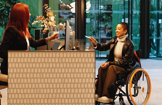 A receptionist seated at a desk in an open office entry reaches for a pass from a woman in a wheelchair.
