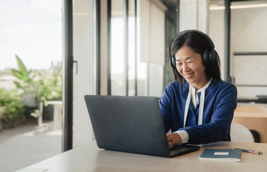 Person wearing headphones and using a laptop at a desk in a bright office, smiling while participating in an online meeting or virtual training session. Large windows and a notebook are visible nearby.