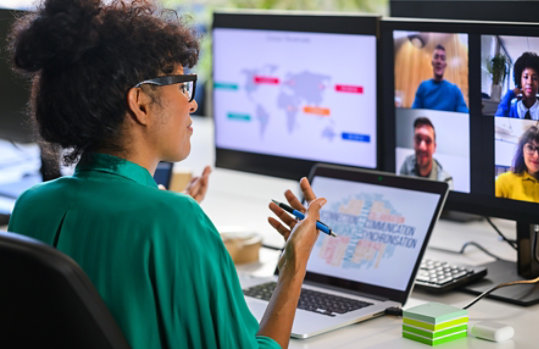 Person participating in a virtual meeting from a desk, speaking while facing multiple screens that display remote colleagues on a video conference call.